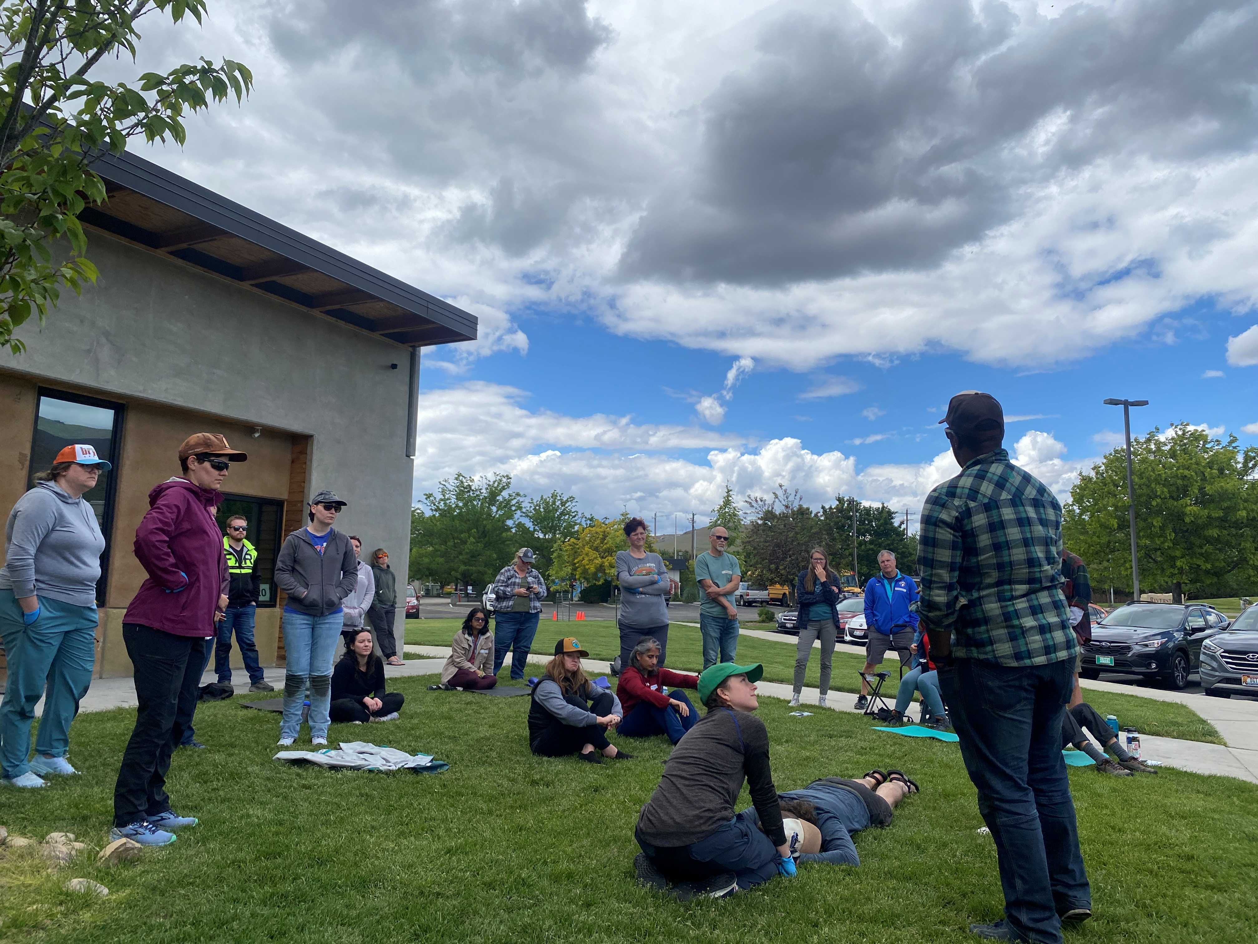 A group of people gather around a demonstration of Wilderness Medicine. Everyone is looking at the instructor while two volunteers get ready to follow his instructions