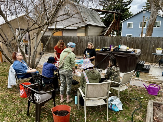 A group of 7 individuals gathered around a table in the outdoors, smiling, planting seeds.
