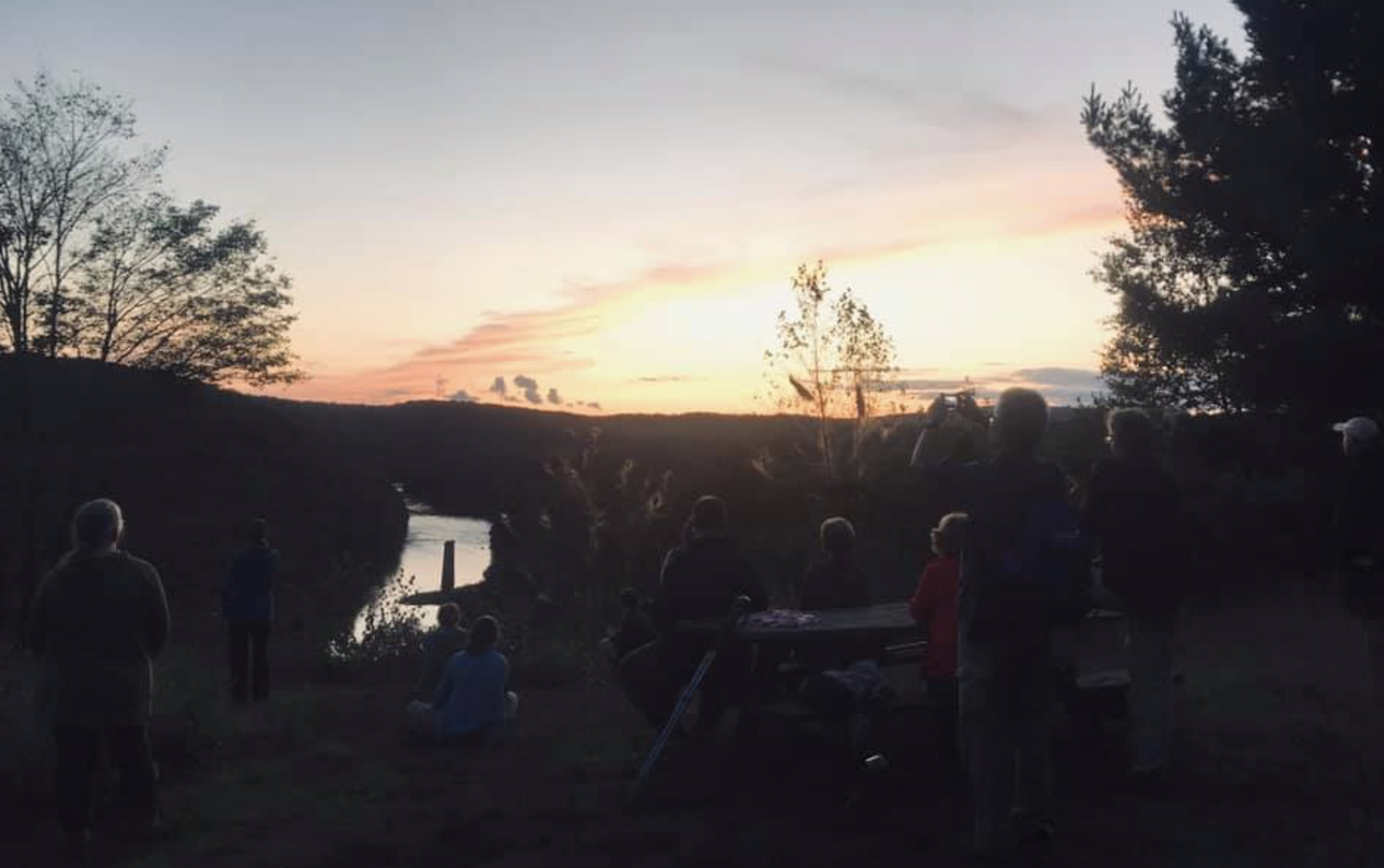 Group of individuals on a cliff at dusk looking at an orange and yellow sunset overlooking a river