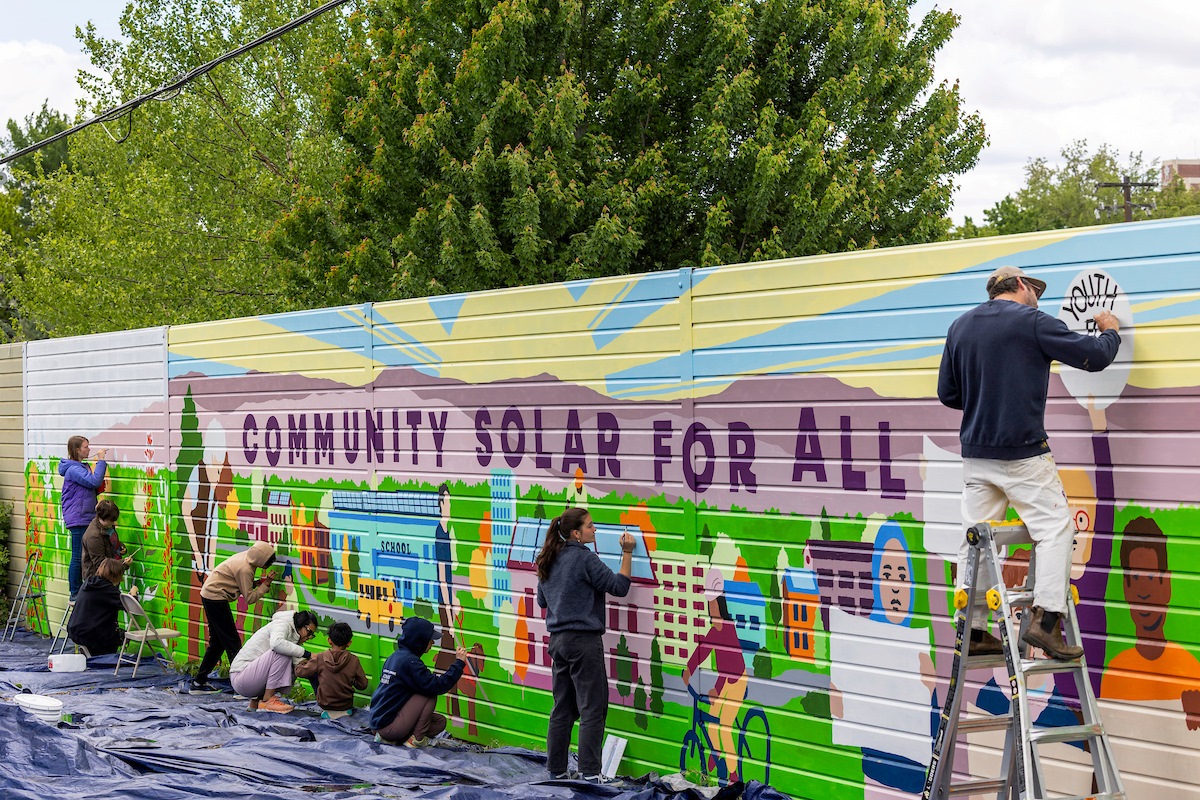 A group of youth and adults paint a mural that says "COMMUNITY SOLAR FOR ALL" with colorful representations of the Boise community.