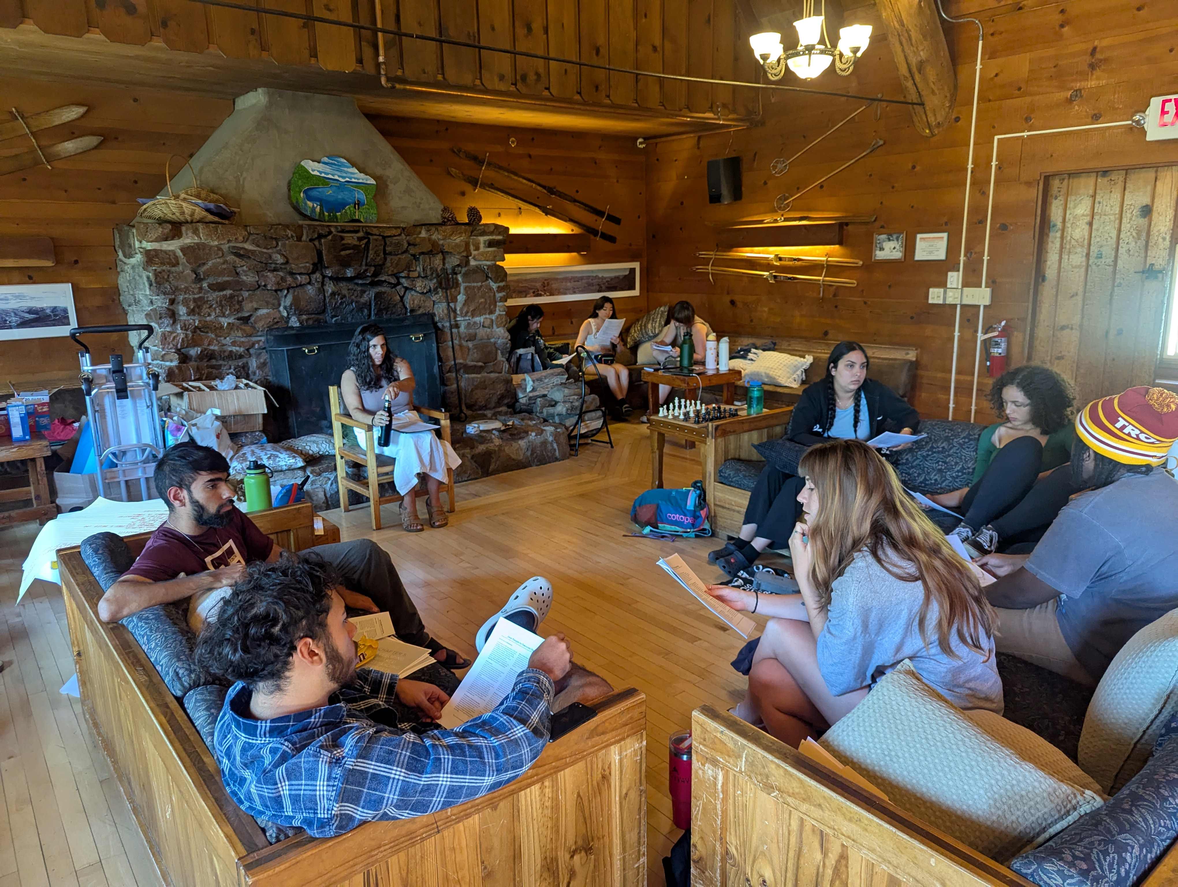 Photo of 5 youth sitting in a circle holding papers and having a discussion during a workshop, in a lodge.