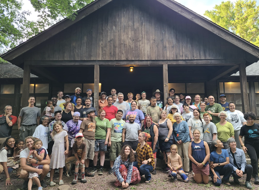 Group photo of many people smiling in front of a wooden cabin