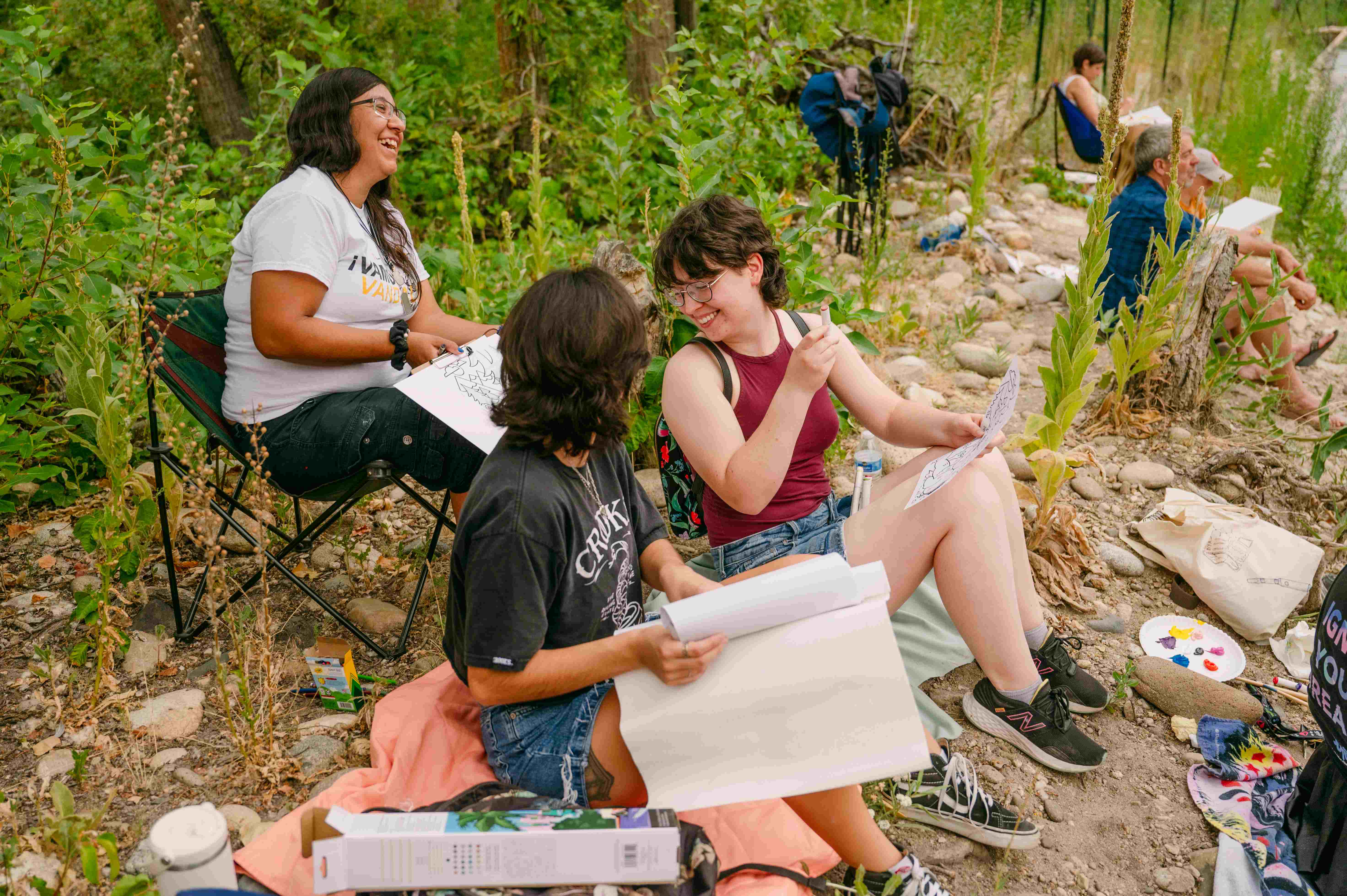 Three individuals sit close to the Boise River, working on various art projects