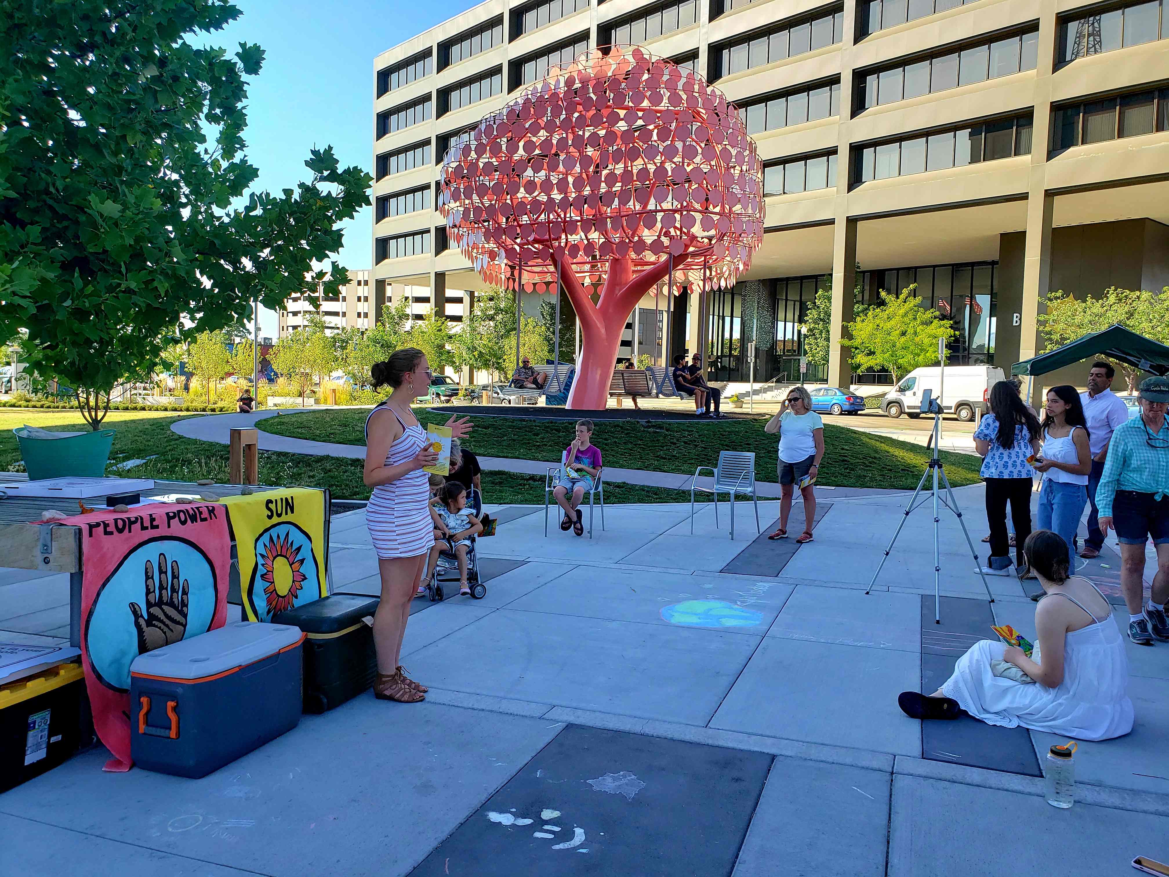 An individual stands in front of a crowd of approximately 10 people giving an MC speech at an urban park with a bright pink tree swing structure