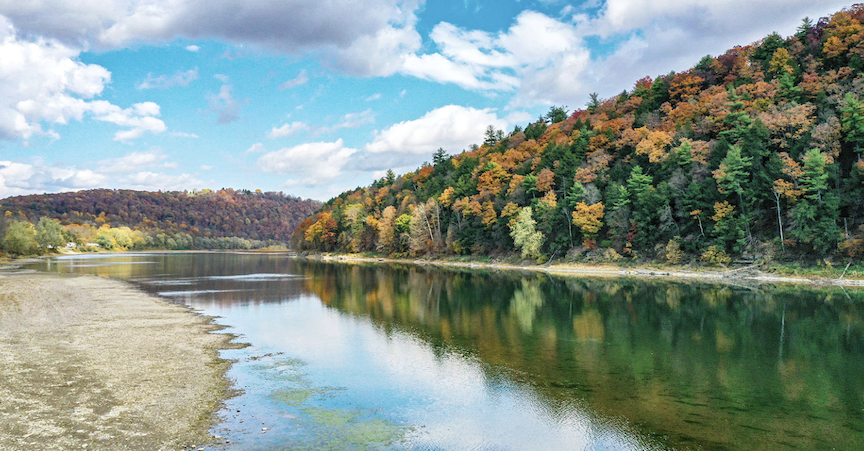 sandy bank on the left with clear sky reflected in calm river water. Fall foliaged trees on right side